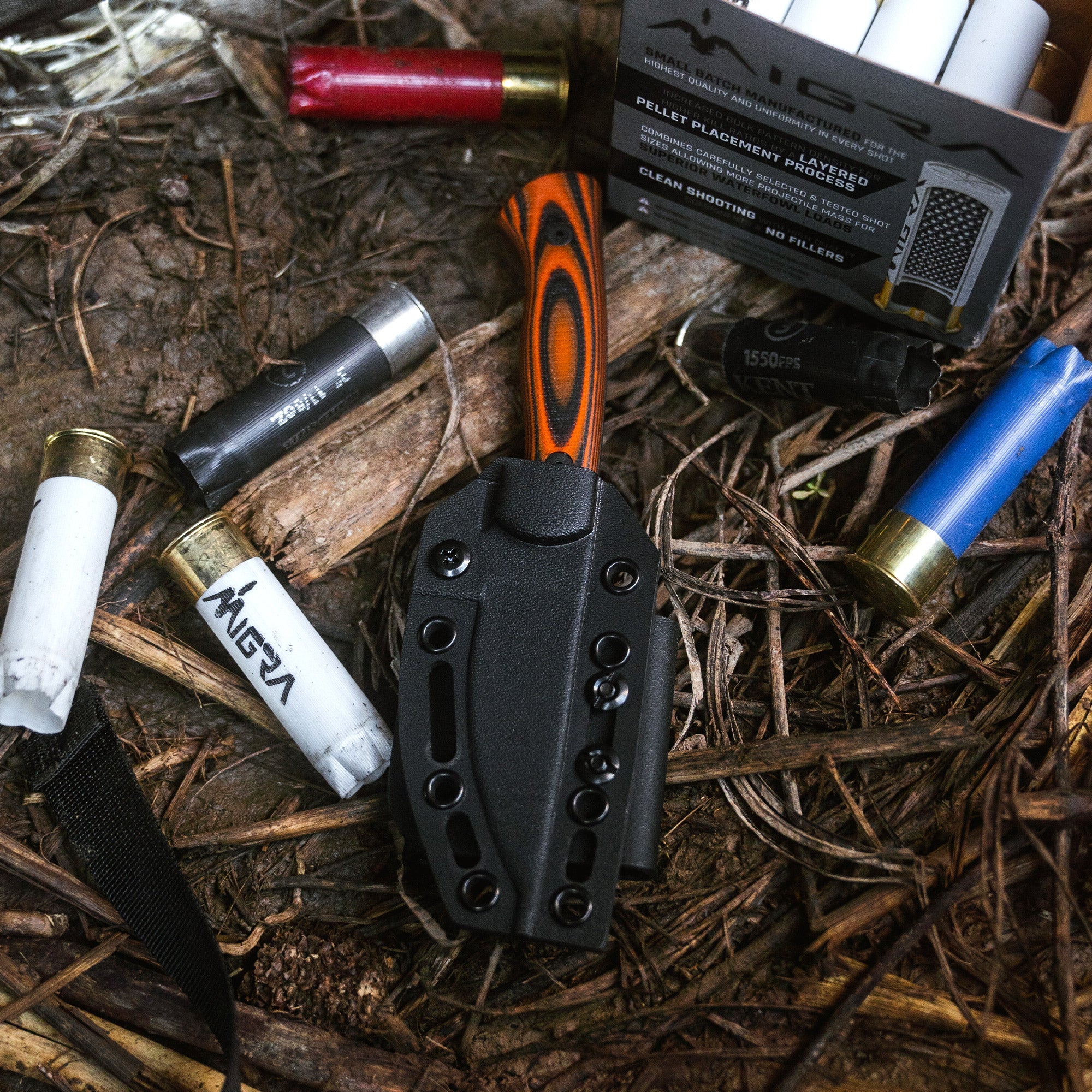 Alt text: "Close-up of a Blackfoot 2.0 additional kydex sheath for a hunting knife, displayed outdoors surrounded by various hunting and outdoor gear including shotgun shells, a flashlight, and a box of pellet placements."