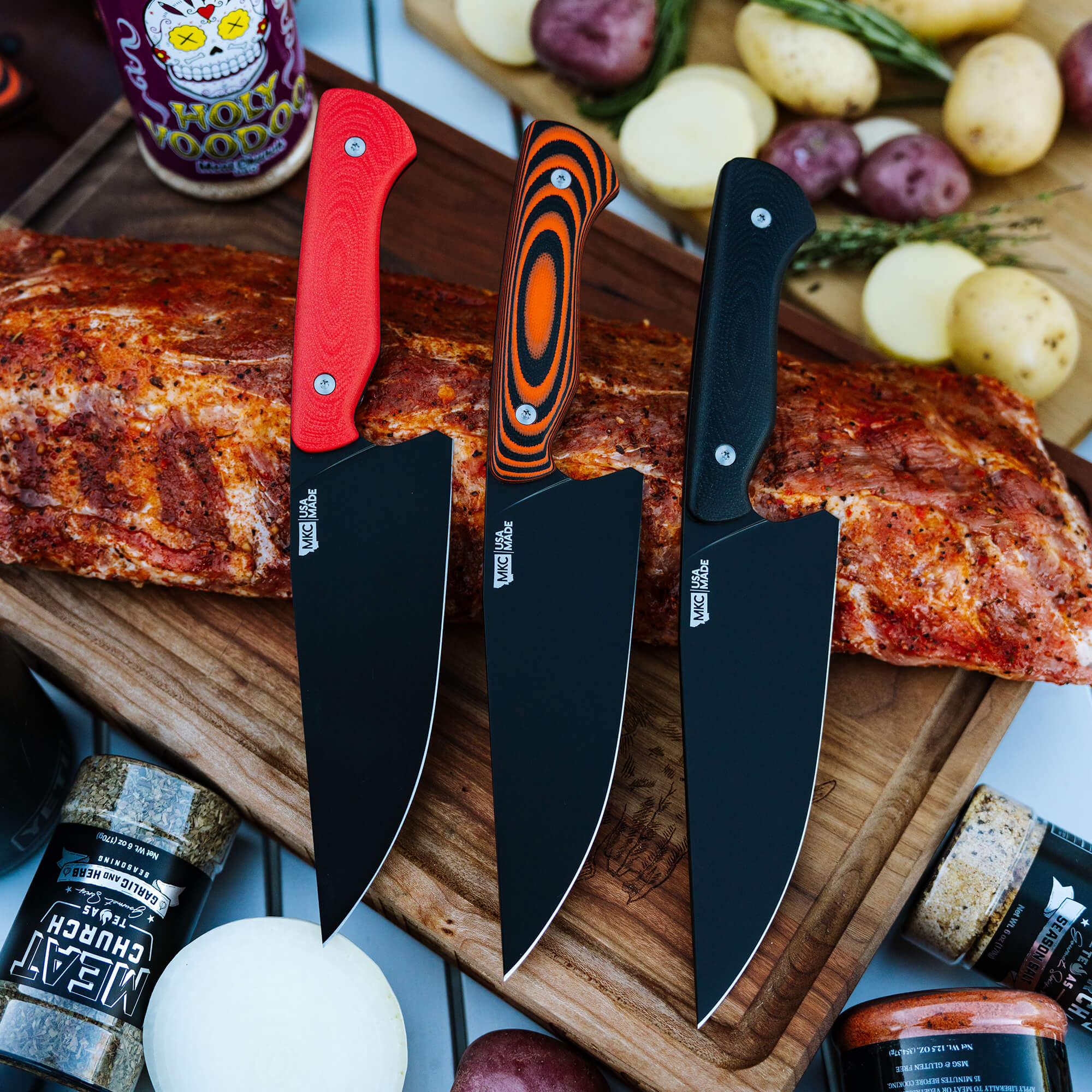 Alt text: Three Meat Church chef knives with distinct red, orange-black, and black handles displayed on a wooden board alongside a cooked rib and various spices.