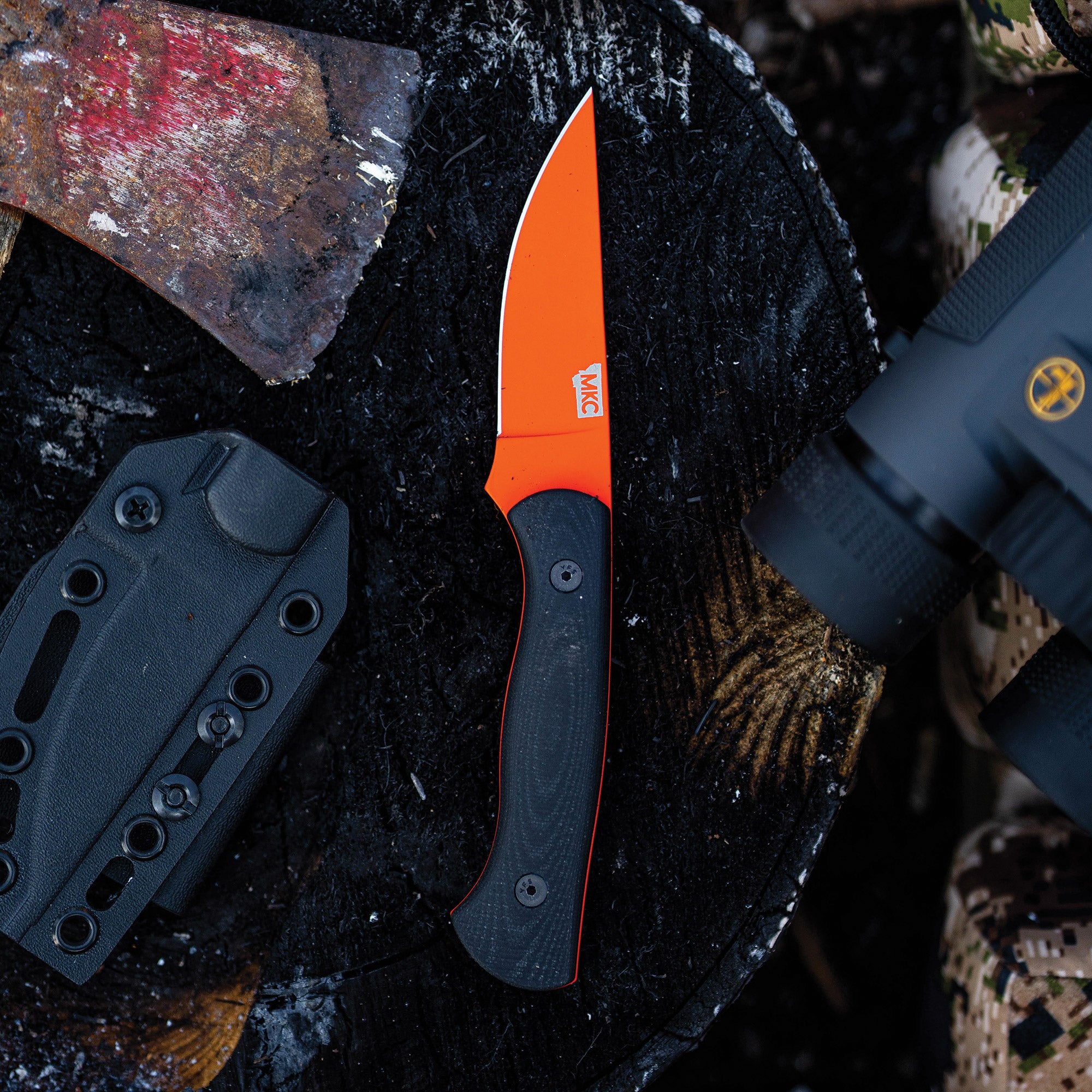 Alt text: "BLACKFOOT 2.0 fixed blade knife with a blaze orange Cerakote™ blade and black handle, displayed alongside a black Kydex sheath, on a burnt wood background with an old rusted axe and part of