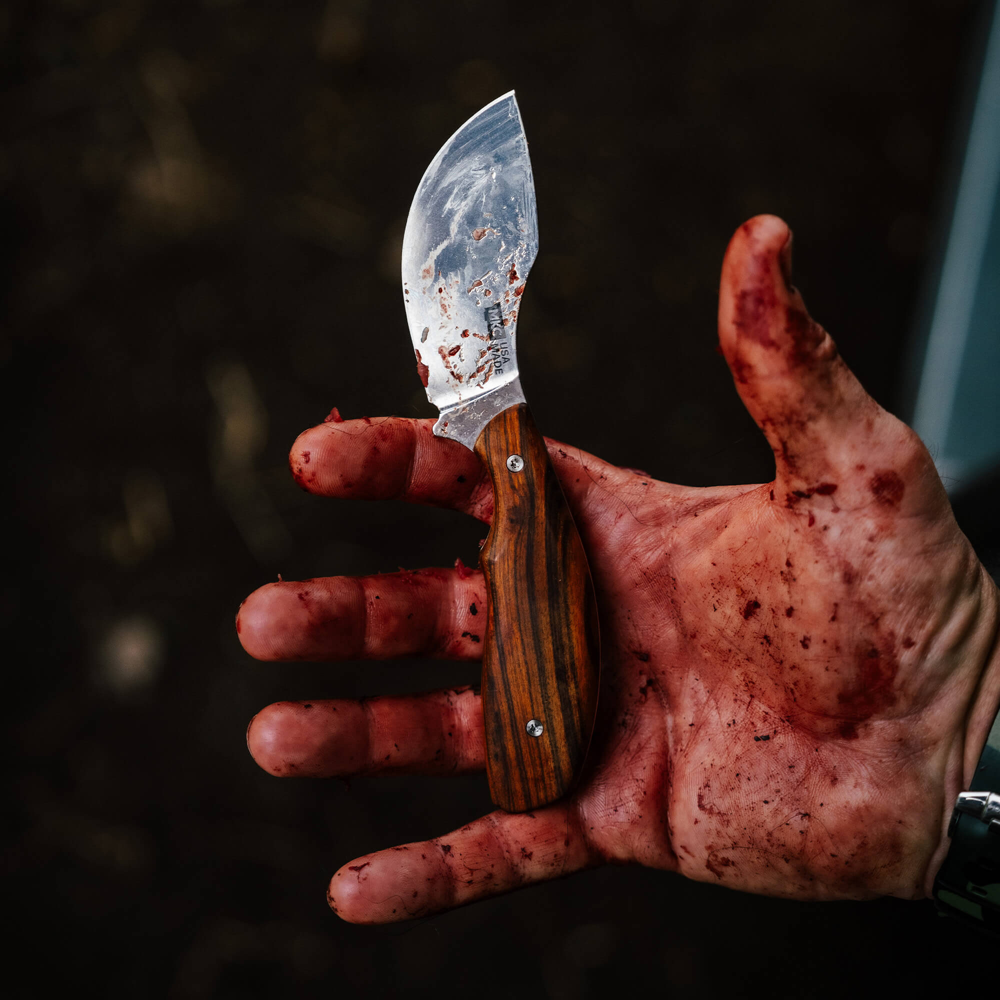KEEP HAMMERING Traditions Packout Skinner knife held in a blood-stained hand, showcasing its curved blade and wooden handle.