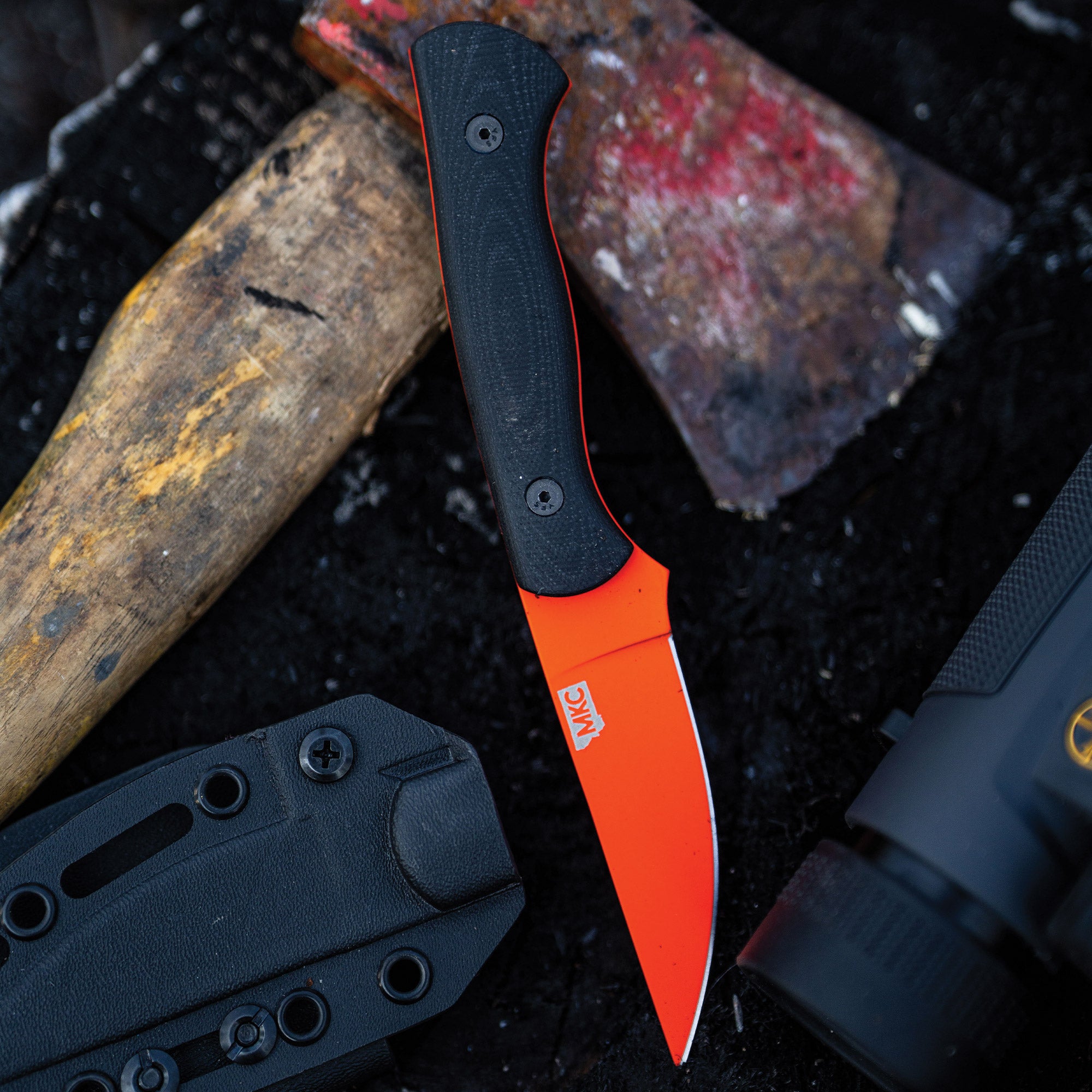 Alt text: "Outdoor scene featuring the BLACKFOOT 2.0 fixed blade knife with a vibrant blaze orange Cerakote coating on the blade and a black handle, displayed next to a black sheath and a worn axe on a gritty,