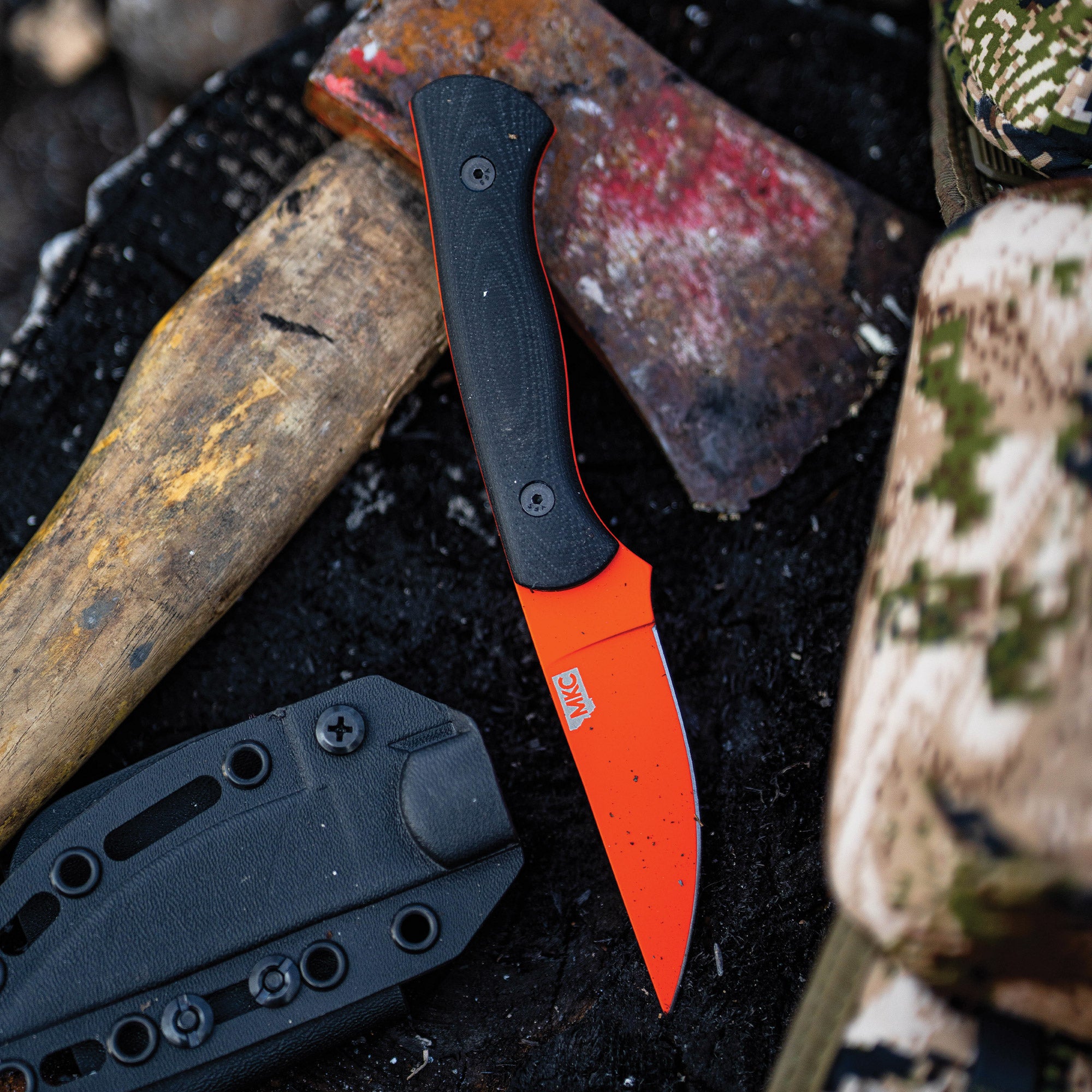 "Blackfoot 2.0 fixed-blade knife with blaze orange Cerakote™ blade surrounded by outdoor gear including a battered hatchet and camouflage items."