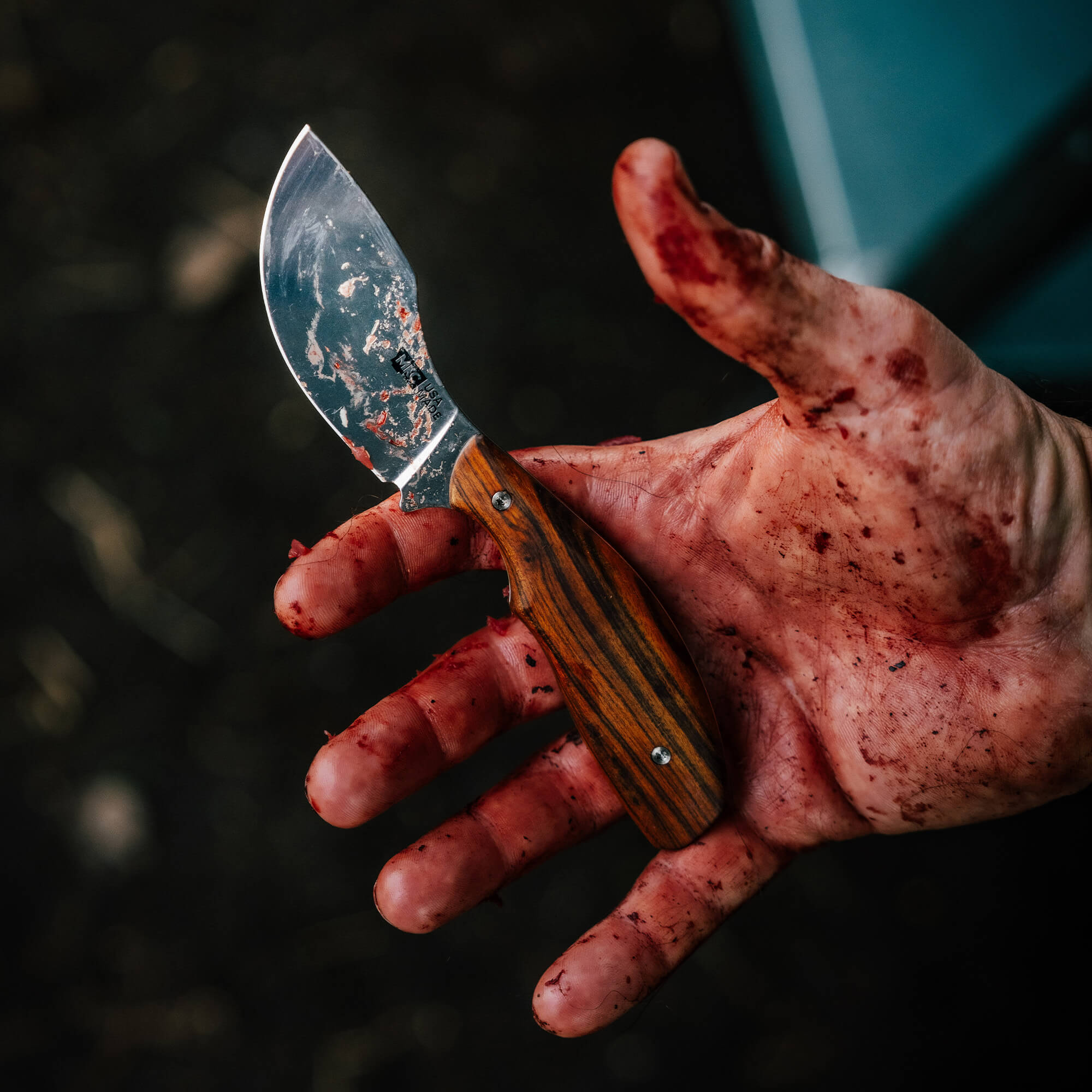 A blood-stained hand holds the KEEP HAMMERING Traditions Packout Skinner knife, showcasing its curved blade and wooden handle.
