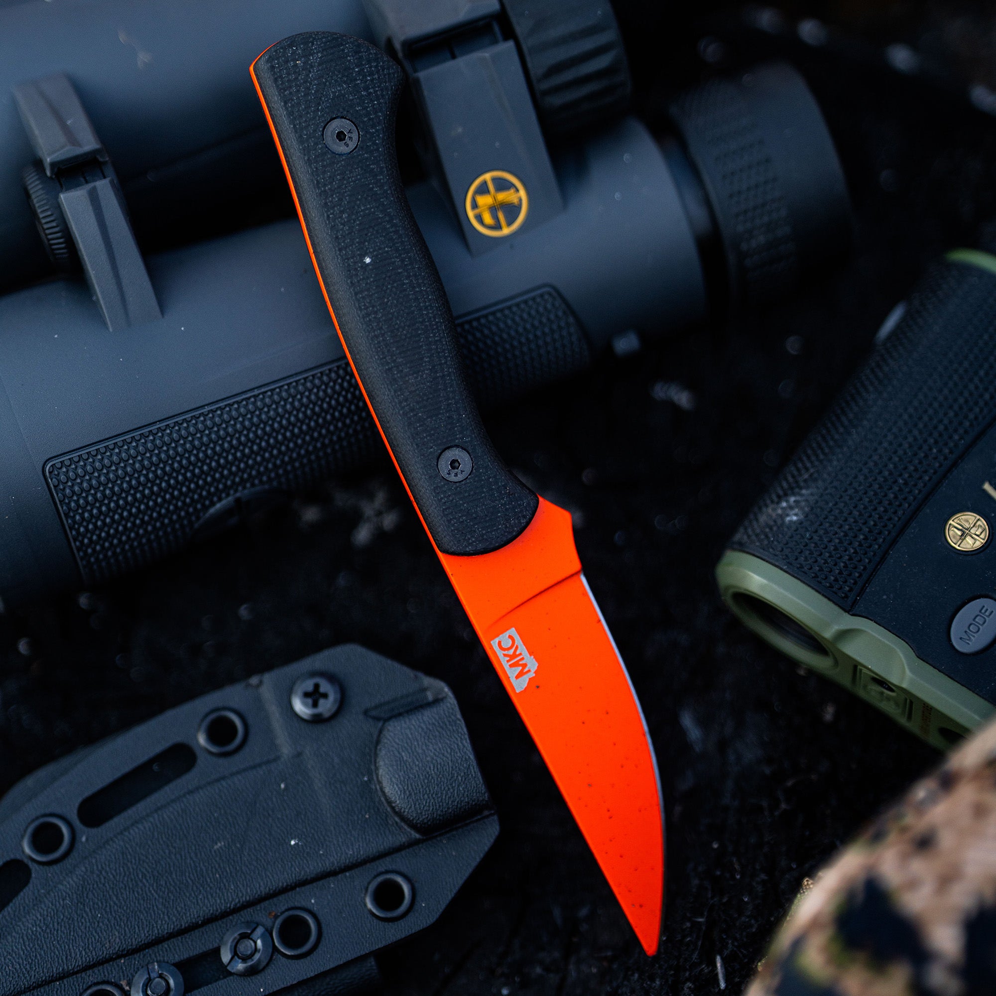 Blaze orange Blackfoot 2.0 knife with black handle accents, displayed alongside tactical equipment, including a black scope and a green textured handgun.