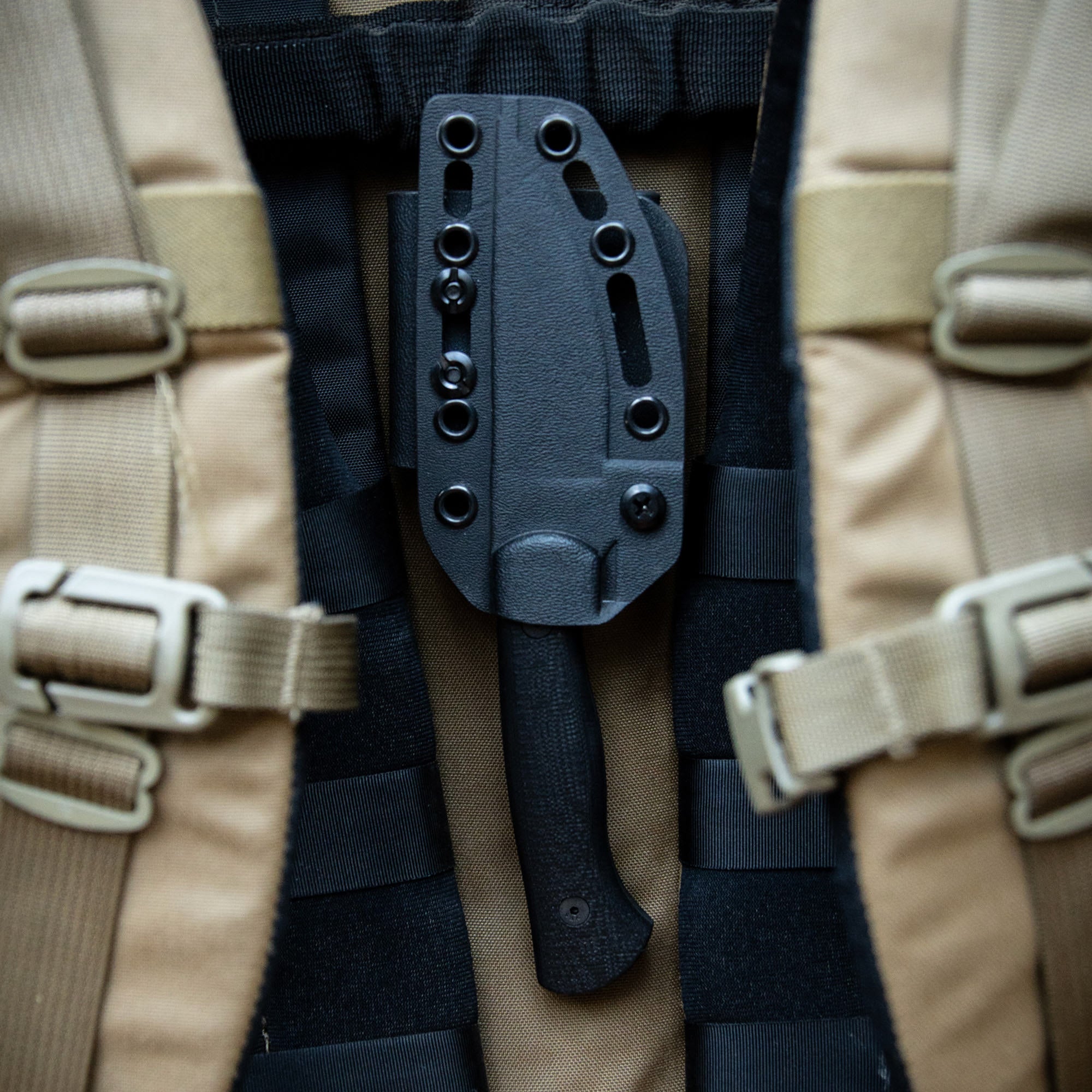 Alt text: "Close-up view of the BLACKFOOT 2.0 tactical knife in a black sheath attached to a military-style vest featuring beige and black stripes with secure fastening."