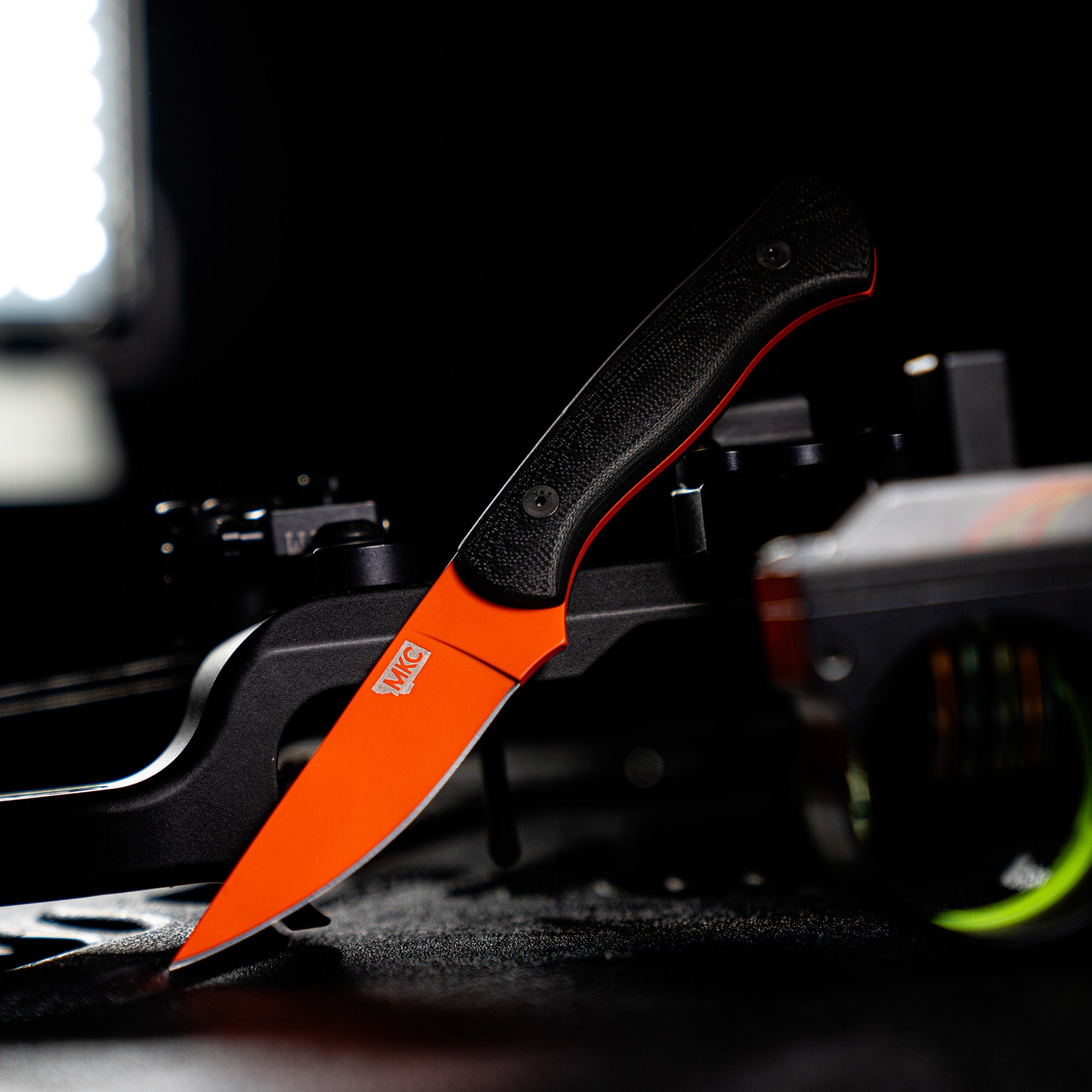 Alt text: Close-up of a Blackfoot 2.0 hunting knife with a blaze orange Cerakote blade and black handle displayed on a dark, textured surface, highlighting its sleek design and the logo on the handle.