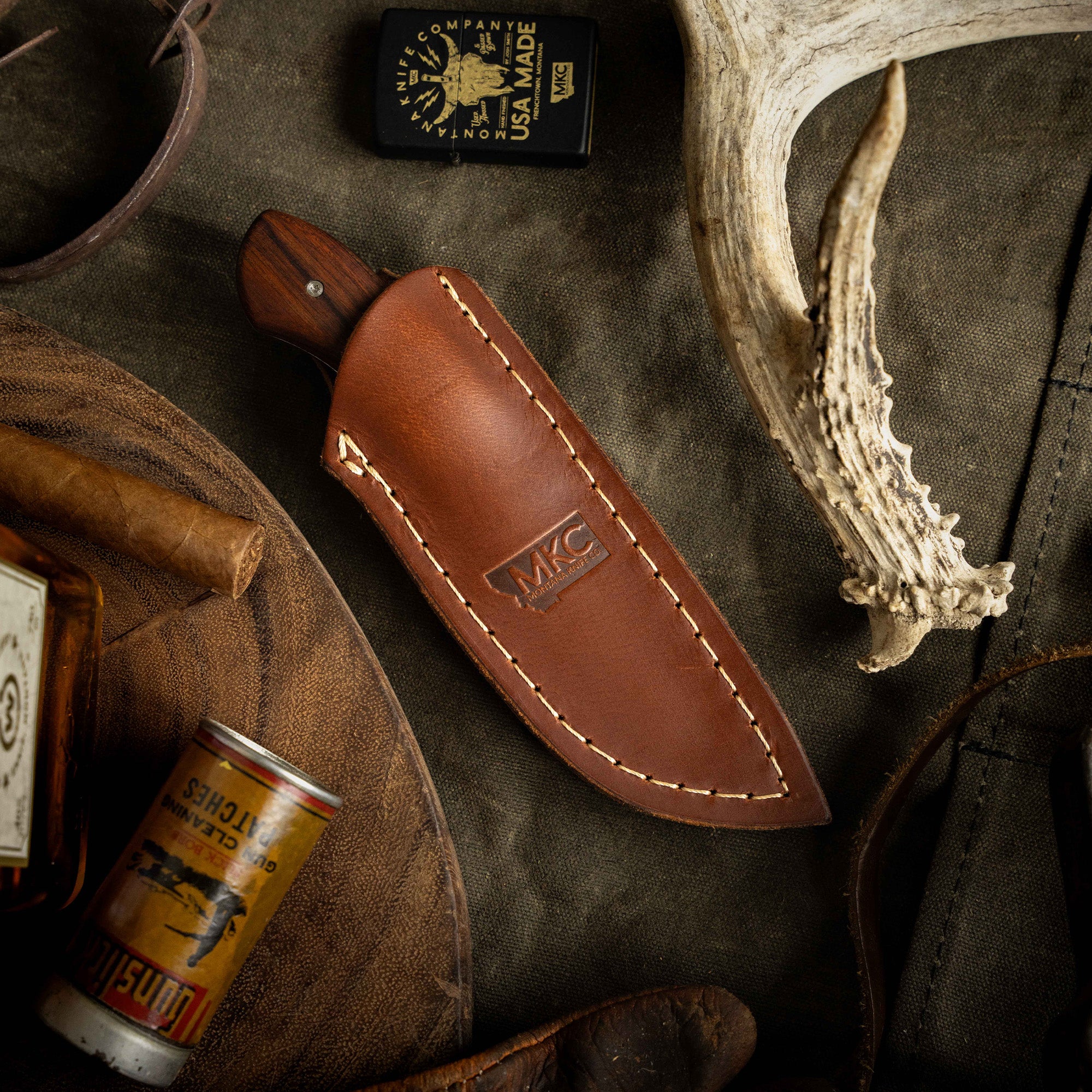 MKC Elk Knife in leather sheath, surrounded by rustic outdoor items, showcasing craftsmanship for hunters and outdoorsmen.