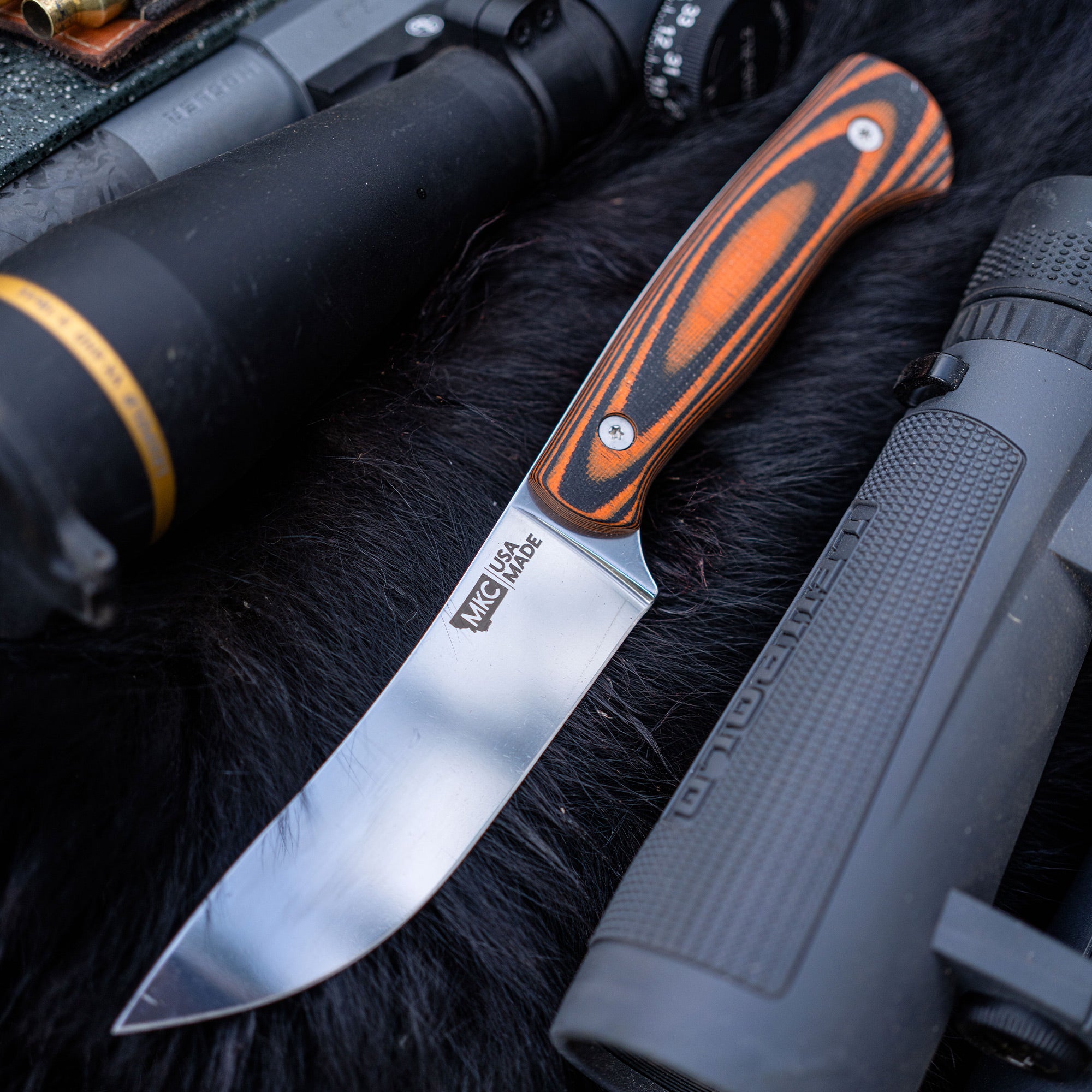 Alt text: "MagnaCut Stonewall Skinner knife with orange and black handle, prominently displaying the sharp, shiny blade, laid on fur next to hunting gear including a black silencer and tactical flashlight."