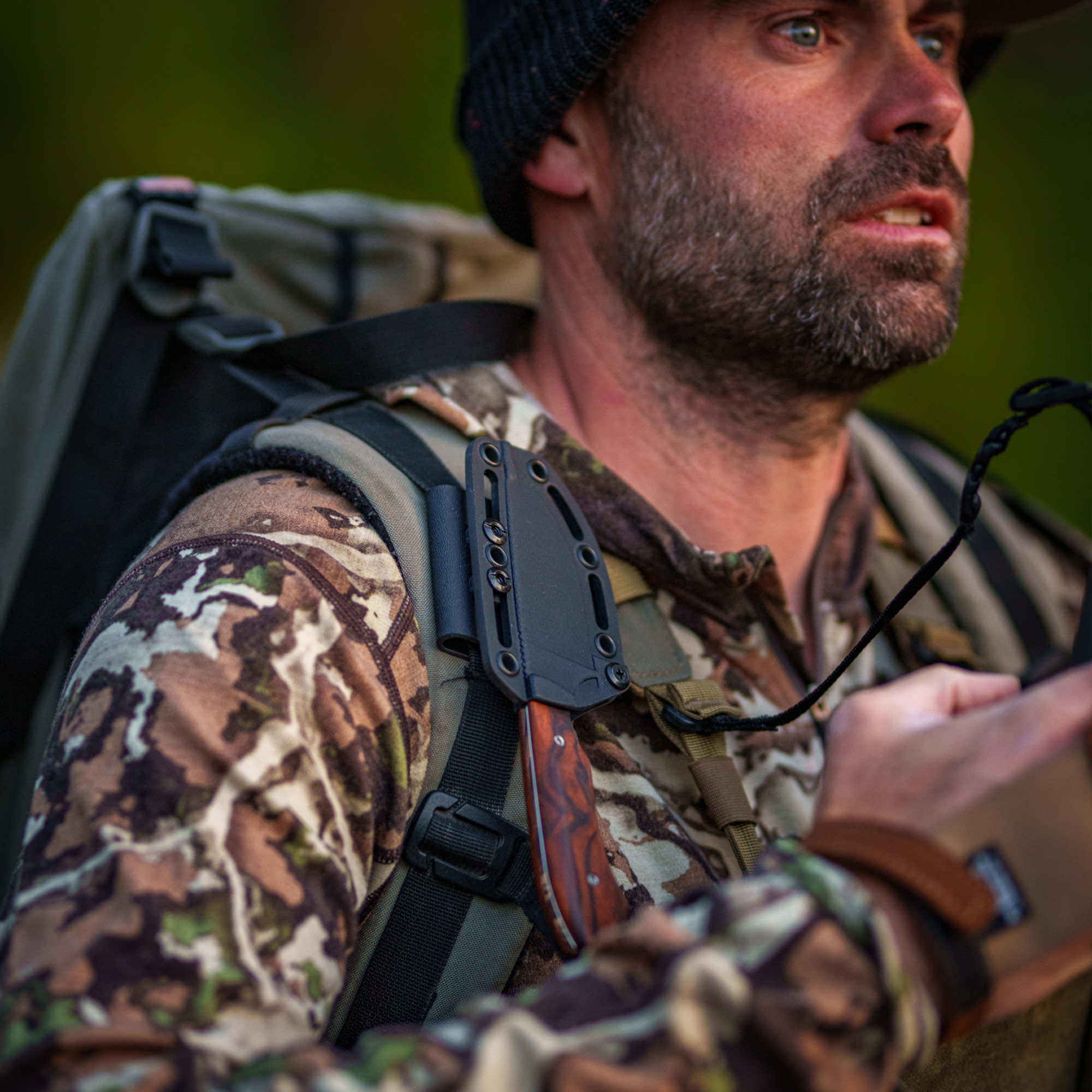 A man in camouflage gear holds a Montana Knife Company elk knife while wearing a backpack in a natural setting.