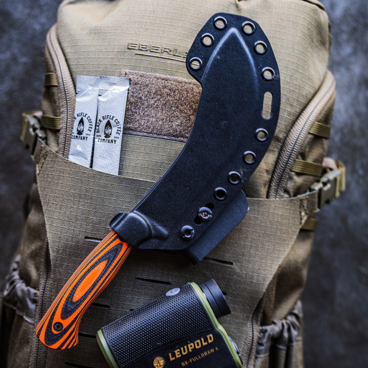 Alt text: Close-up of a black Kydex sheath for a knife with a vibrant orange handle, attached to a tactical backpack displaying the Eberlestock logo, alongside outdoor gear including a Leupold binocular.