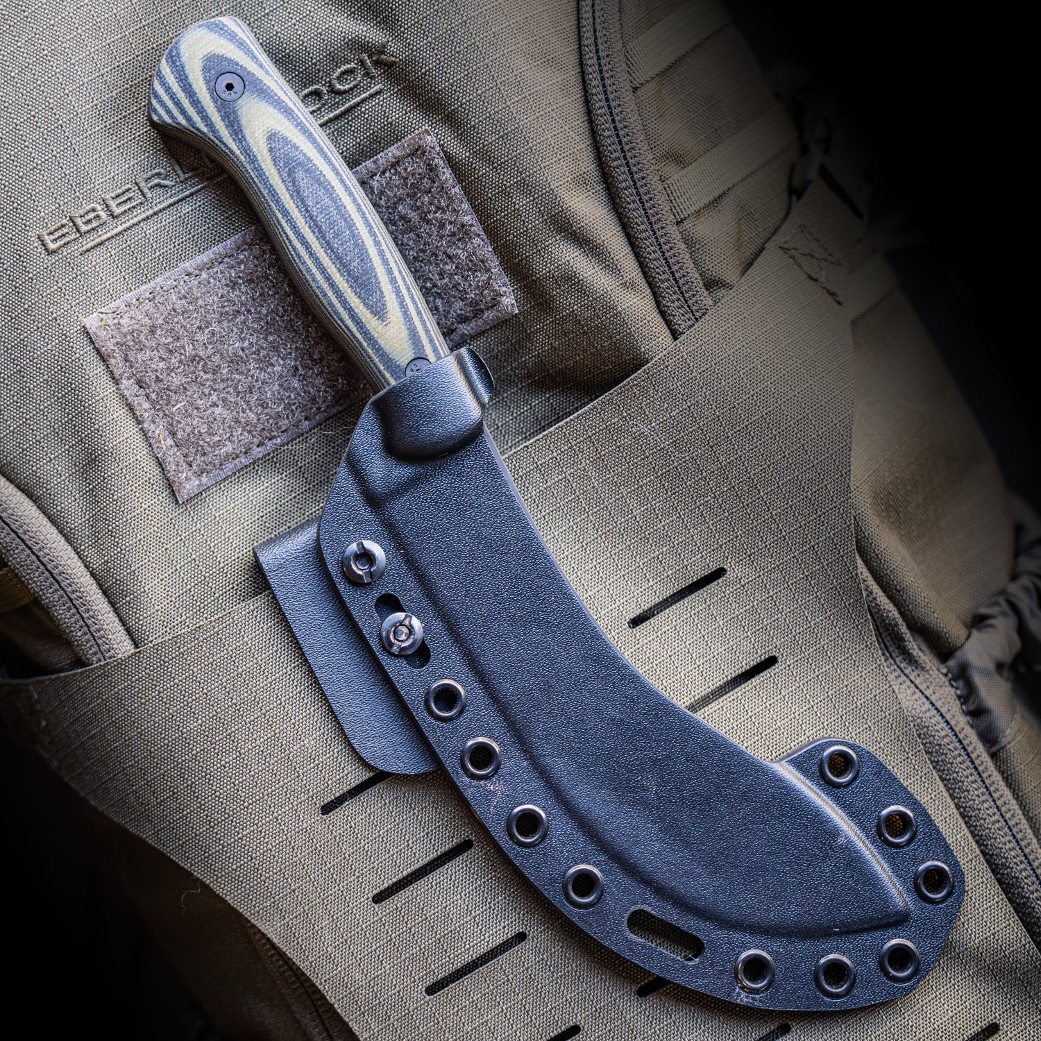 Alt text: "Close-up of a Beartooth Pro Skinning Blade with a tan and black handle partially sheathed in a black Kydex sheath, mounted on a beige tactical vest."