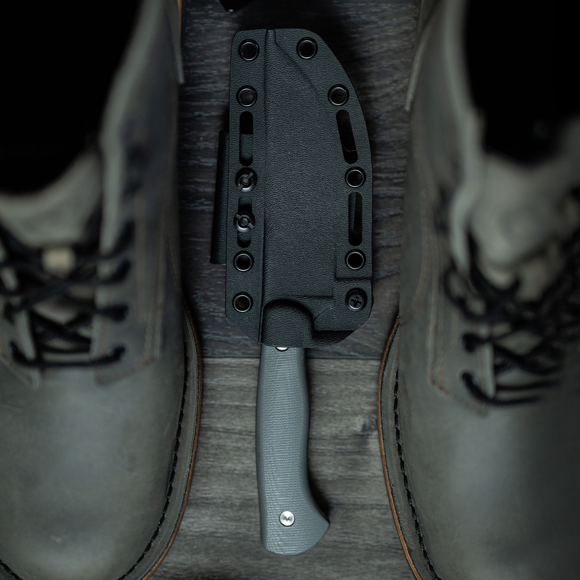 Alt text: "Magnacut Stonewall Skinner knife with a gray handle, showcased between two dark leather boots, encased in a black sheath designed for attachment, displayed on a wooden background."