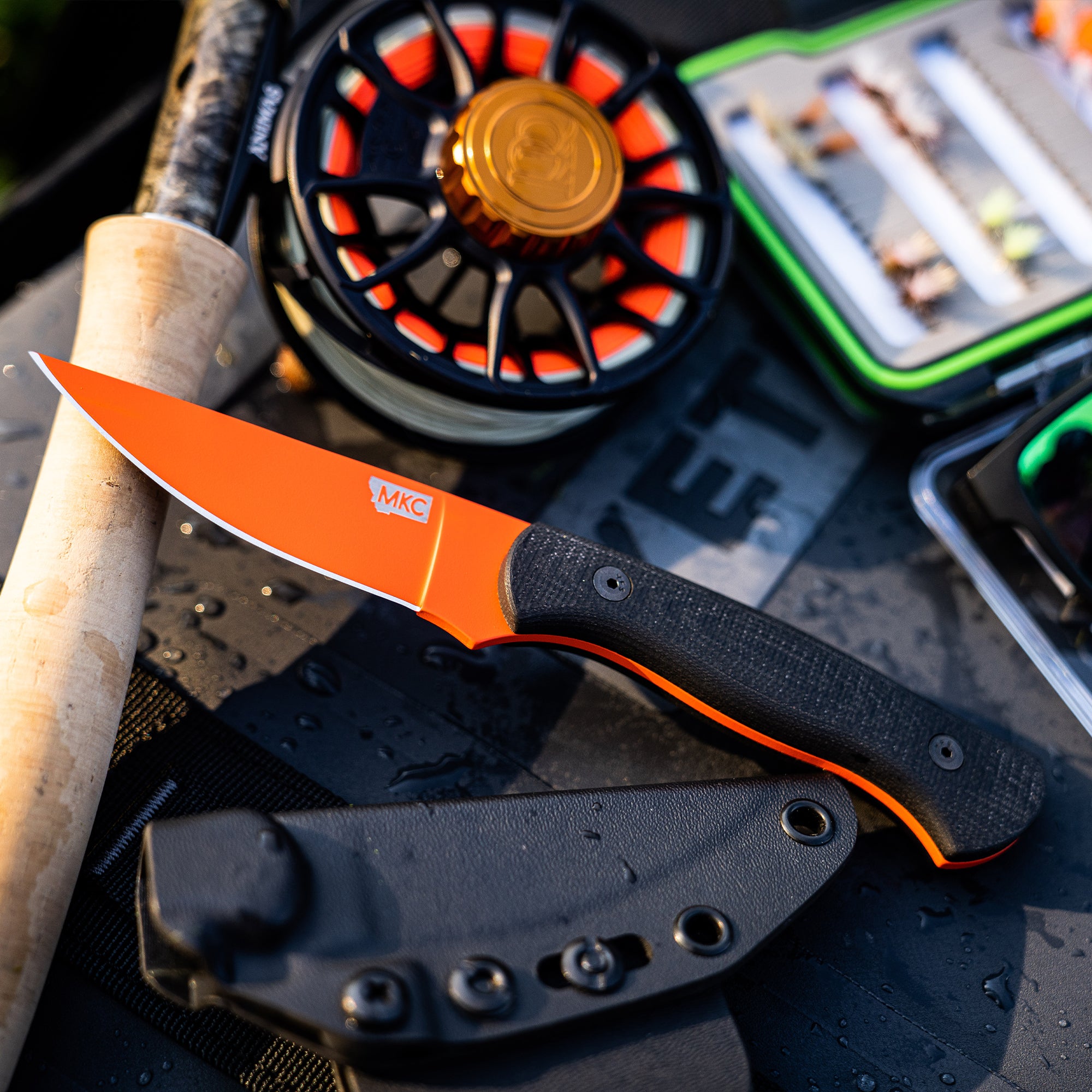 "Close-up of the BLACKFOOT 2.0 knife with a blaze orange Cerakote blade, displayed alongside a fishing rod, reel, and tackle box on a wet surface, highlighting outdoor utility and durability."