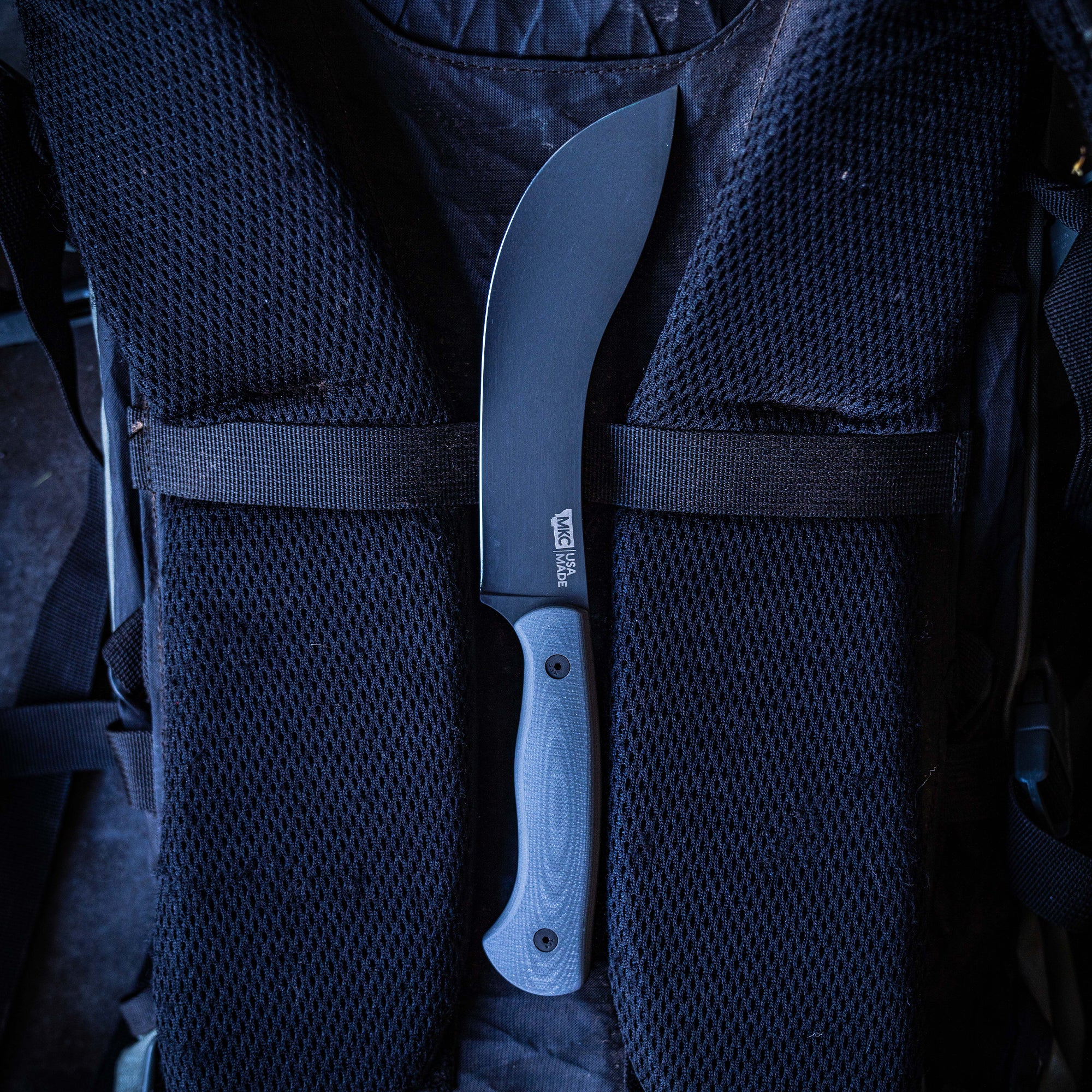 Alt text: Grey Beartooth Pro Skinning blade with a curved edge and ergonomic light grey handle securely placed in a woven black mesh backpack strap.