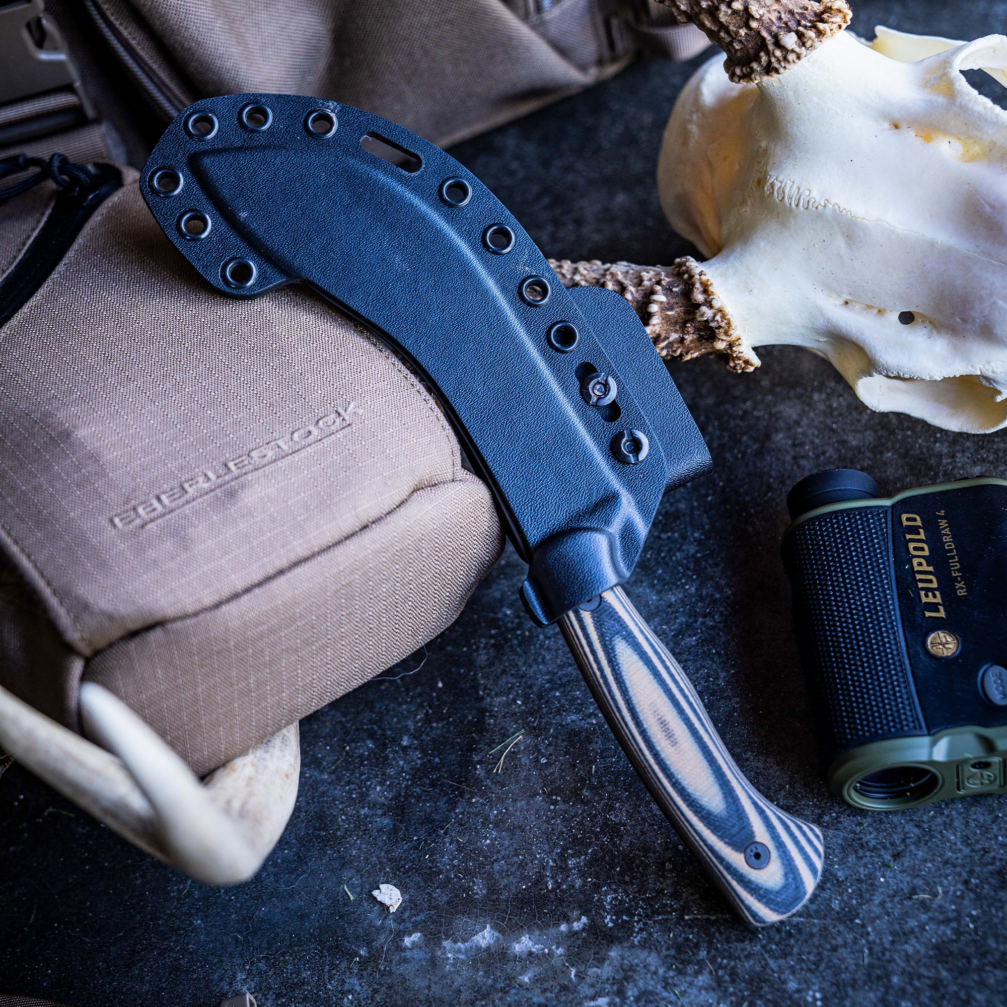Alt-text: "BEARTOOTH Pro Skinning Blade with tan and black handle laid next to its black sheath, beside a beige tactical bag and a skull, with a Leupold monocular in the background, displayed on a textured dark